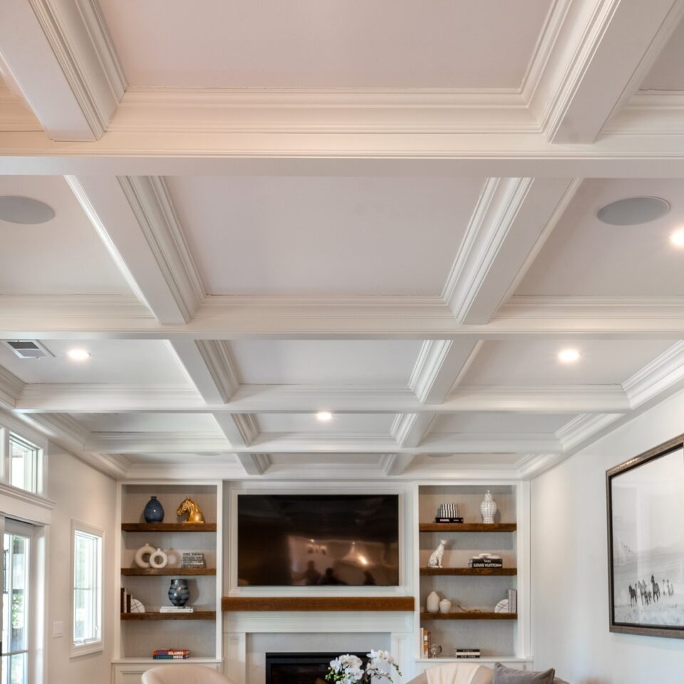 Elegant living room with coffered ceiling, built-in shelving, modern fireplace, and neutral-toned furniture arranged around a beautiful l display.