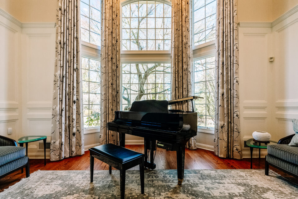 Grand piano positioned in front of tall arched windows with custom drapery, surrounded by tailored seating and decorative accents in a sophisticated music room.